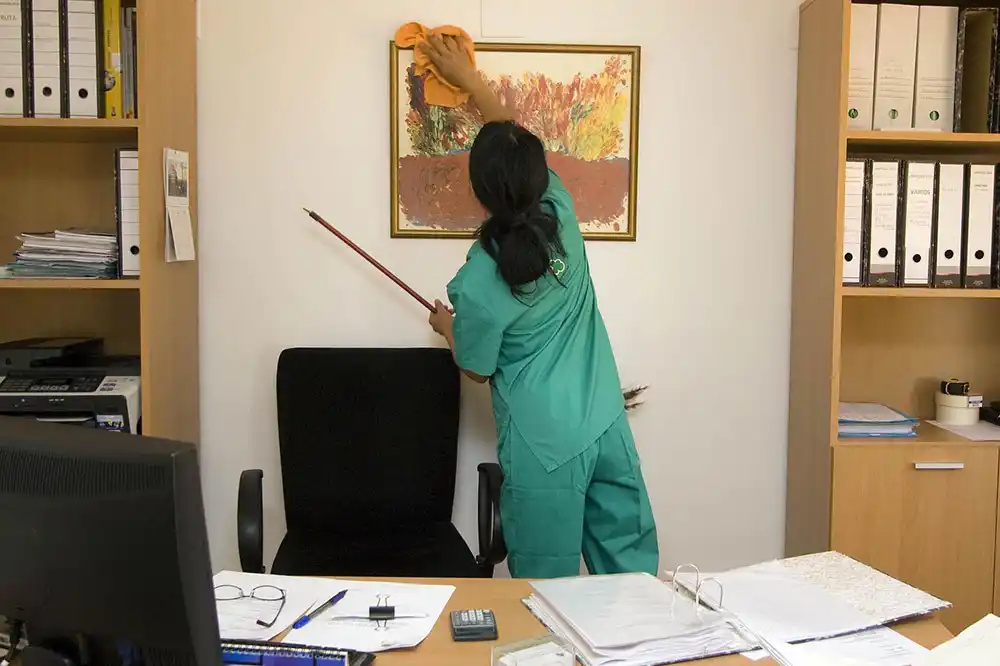 Woman janitor using dust cloth to clean the top of a picture in an office