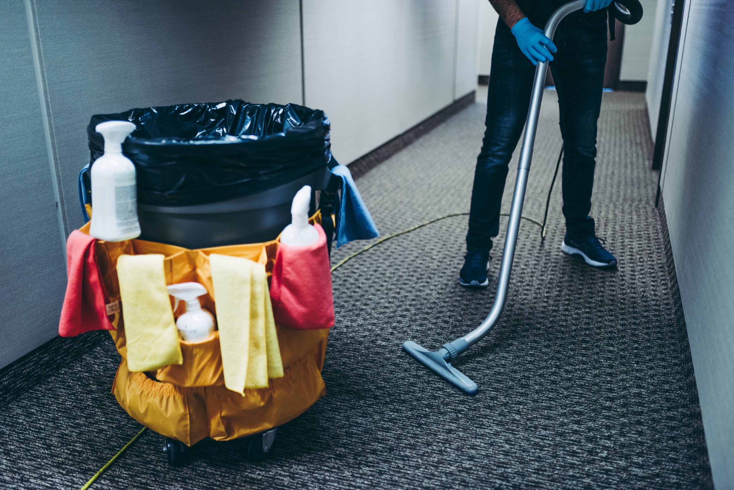 Trash can with cleaning supplies next to janitor vacuuming floor