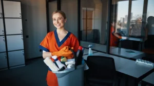 Woman janitor in orange uniform standing in office with cleaning bucket