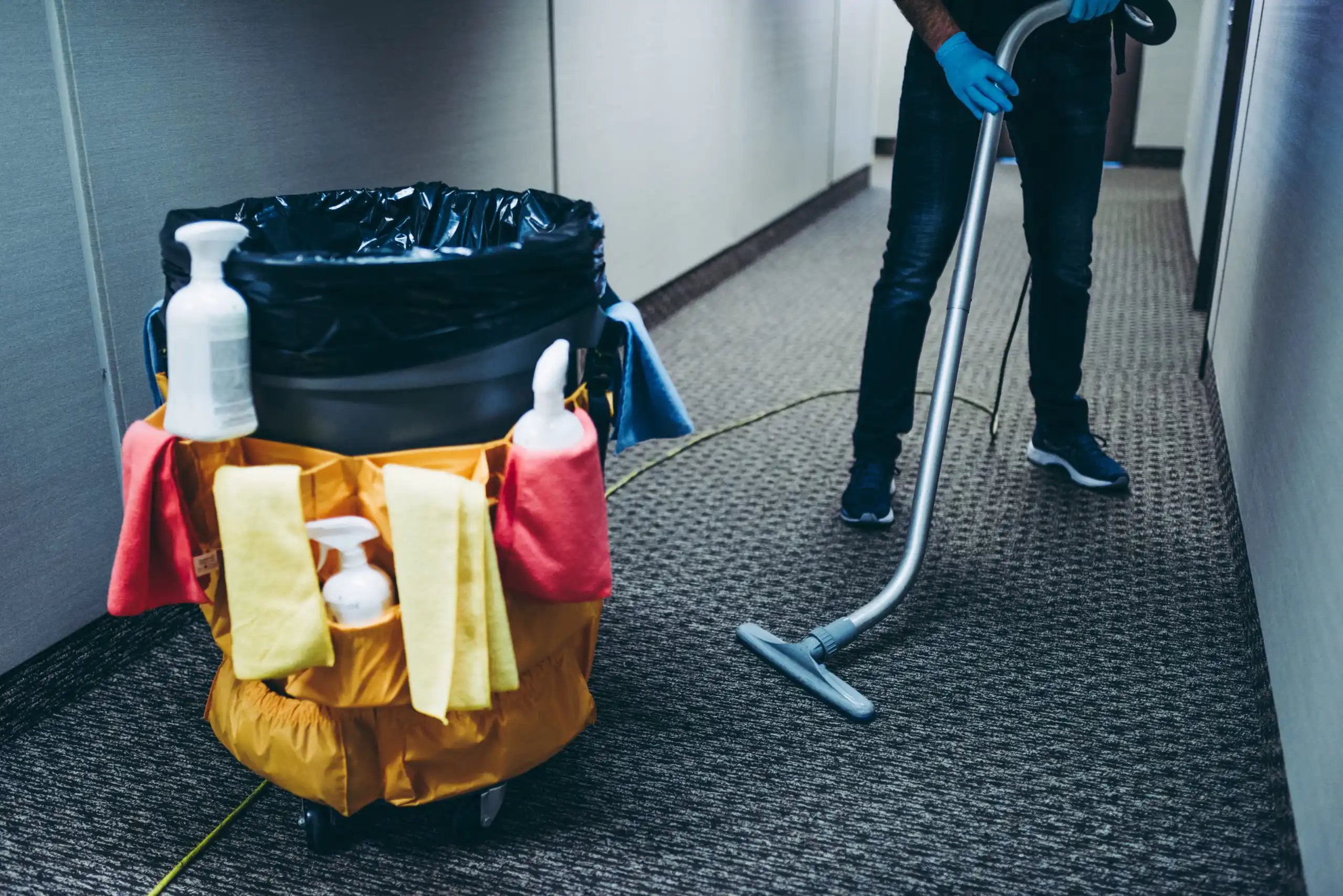 Janitor vacuuming and cleaning around trash can with cleaning supplies