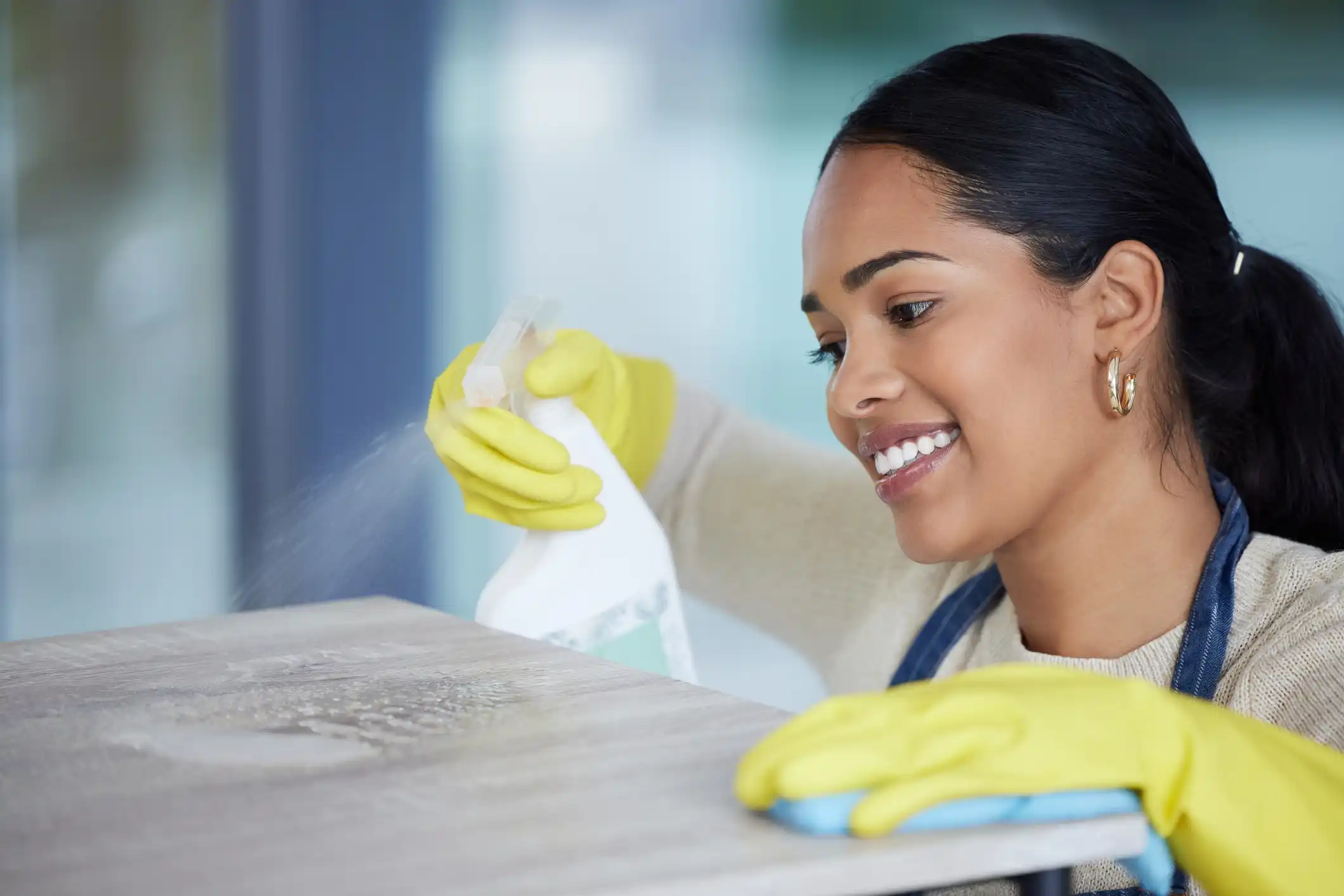 Woman smiling with apron on cleaning counter with yellow gloves , spray and sponge