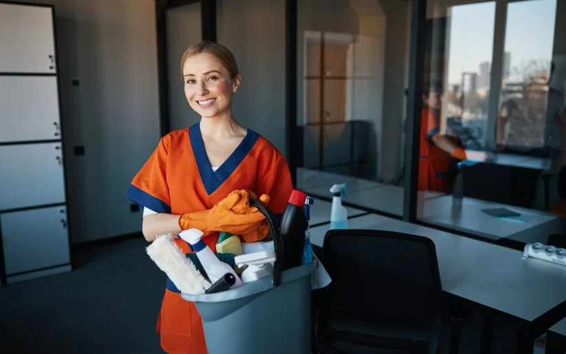 Woman janitor in orange uniform standing in office with cleaning bucket